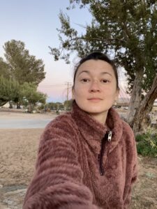 A selfie of Becca in a fuzzy pink sweater, the desert sand and sky behind her.